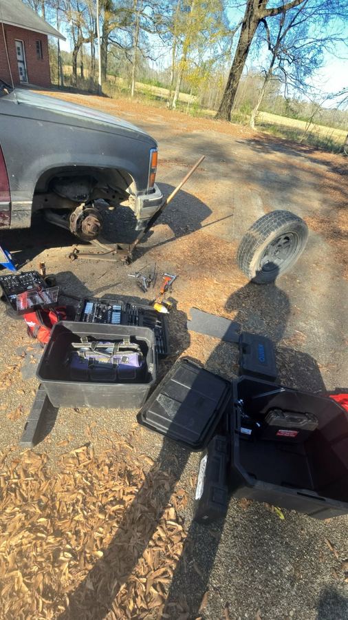 Pickup truck raised on a jack in a gravel driveway with tools laid out for a wheel-off repair